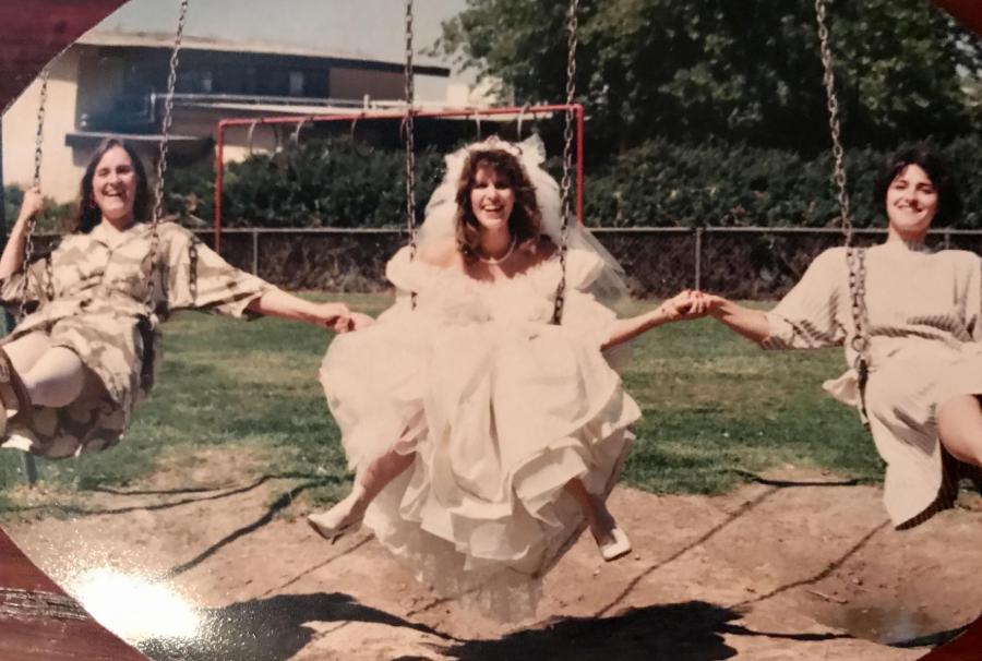 Three women, including a bride in a white dress and veil, are sitting on swings outdoors, holding hands and smiling. Two women in dresses sit on either side of the bride. Trees and playground equipment are in the background.