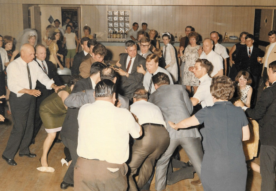 A large group of people in formal attire are gathered in a wood-paneled room, appearing to be involved in a lively or chaotic activity, with some people laughing and others watching closely.