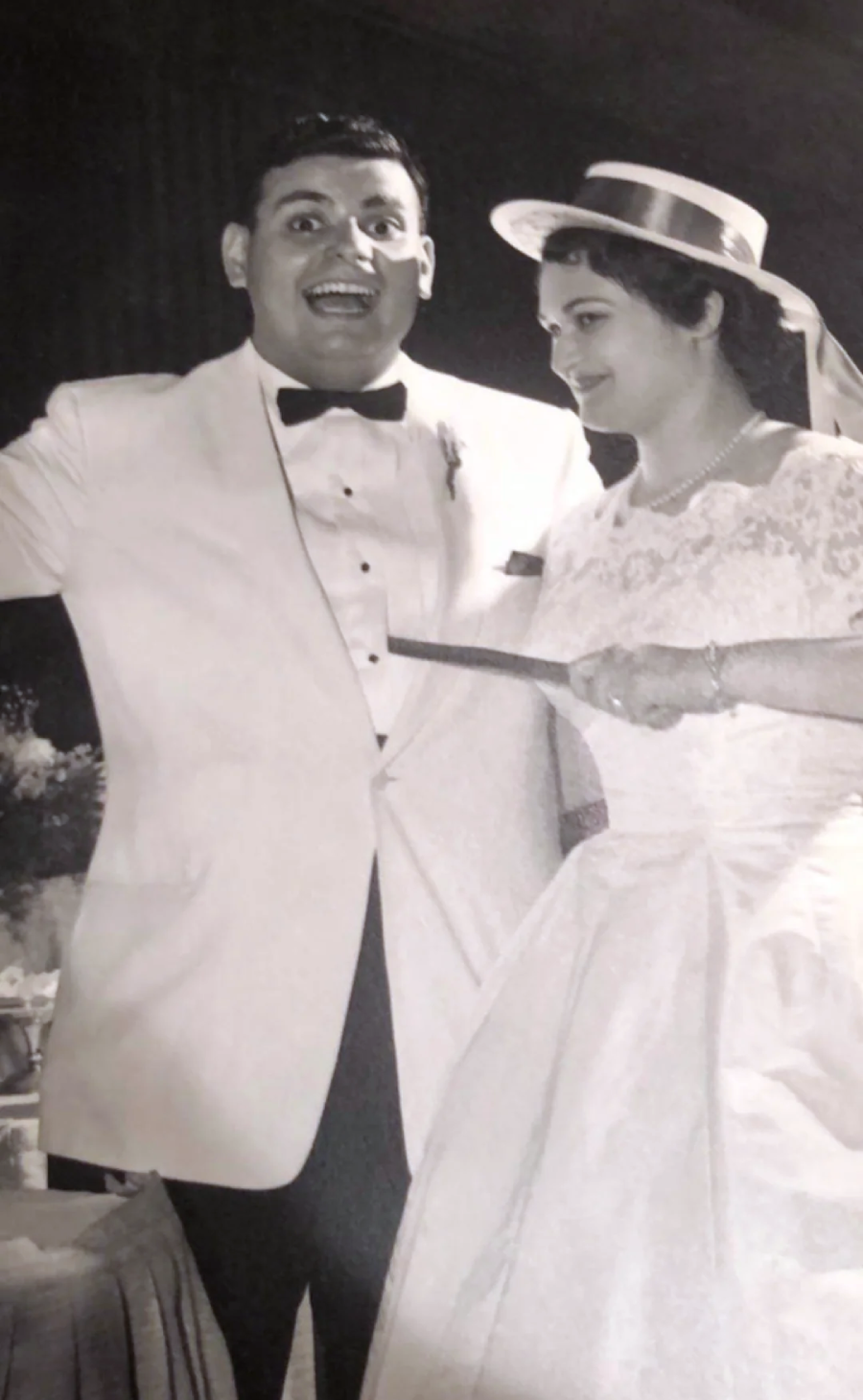 A joyful bride and groom in formal wedding attire, smiling as they cut their wedding cake together. The groom wears a white tuxedo and bow tie; the bride wears a lace wedding dress and veil with a hat. Black and white photo.