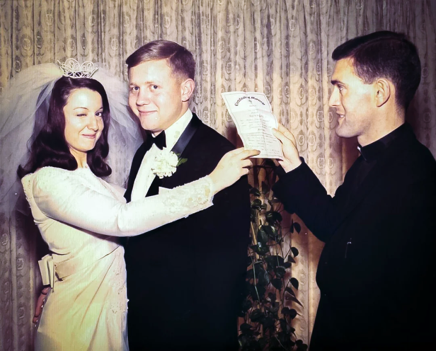 A bride and groom stand together, smiling, with the bride pointing at a document held by a priest. They are dressed in wedding attire, in front of a curtain backdrop with a leafy plant behind them.