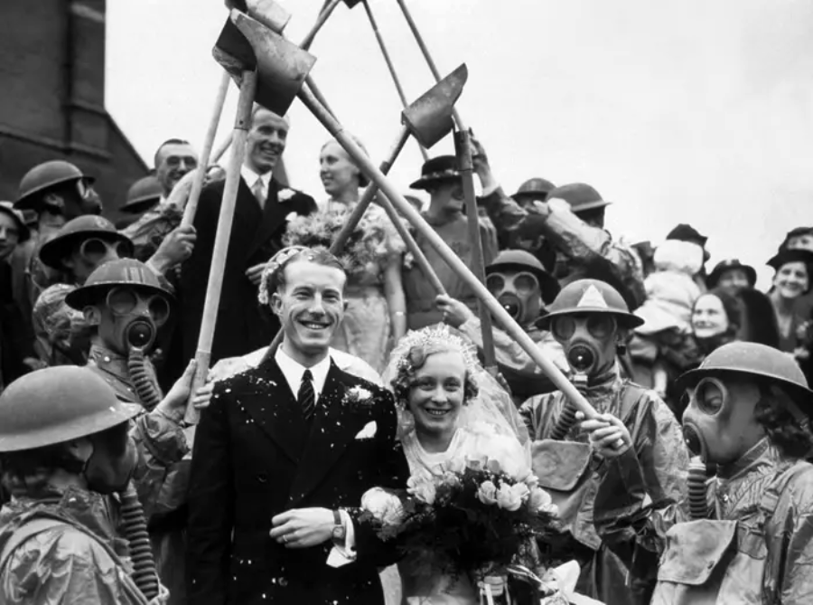 A bride and groom smile as they exit a building, surrounded by people in gas masks and helmets raising axes overhead, with confetti falling and a crowd celebrating in the background.