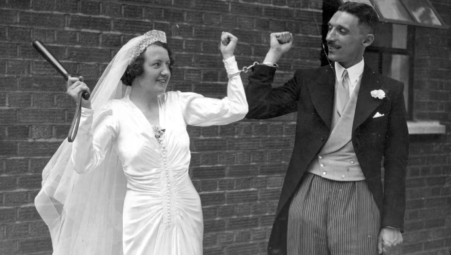 A smiling bride in a white wedding dress and veil holds a baton and is handcuffed to a groom in a suit. Both raise their arms playfully, standing in front of a brick wall.