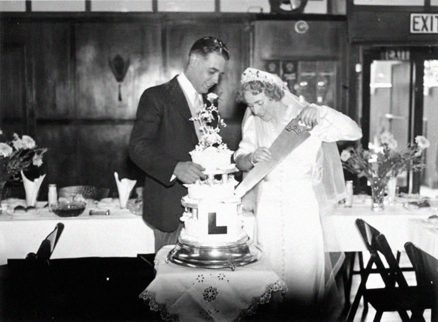 A bride and groom stand together cutting a tiered wedding cake with a large saw at a decorated reception table, surrounded by flowers and table settings.