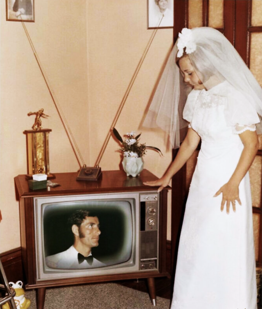 A bride in a white dress and veil stands beside a vintage TV showing a man in a tuxedo. The TV is topped with a small vase of flowers and a trophy, and the room has framed photos and tan walls.