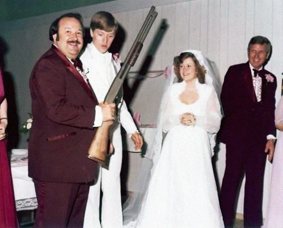 A man in a maroon suit holds a shotgun and smiles next to a nervous groom in white, a smiling bride in a white dress, and another man in a maroon suit, all standing indoors at a wedding.