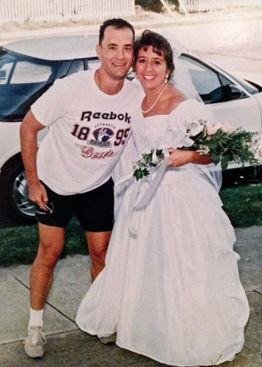 A smiling bride in a white wedding dress stands next to a man in a casual Reebok t-shirt and shorts. They pose together outdoors near a white car, with greenery and pavement in the background.