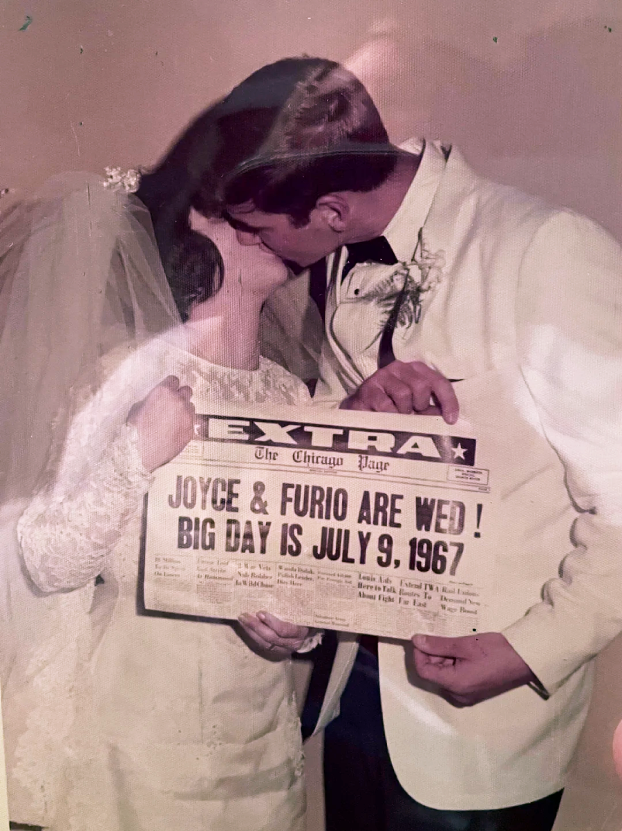 A bride and groom in wedding attire kiss while holding a newspaper-style sign that reads, "Joyce & Furio are wed! Big day is July 9, 1967." The bride wears a veil and lace dress; the groom wears a white suit.