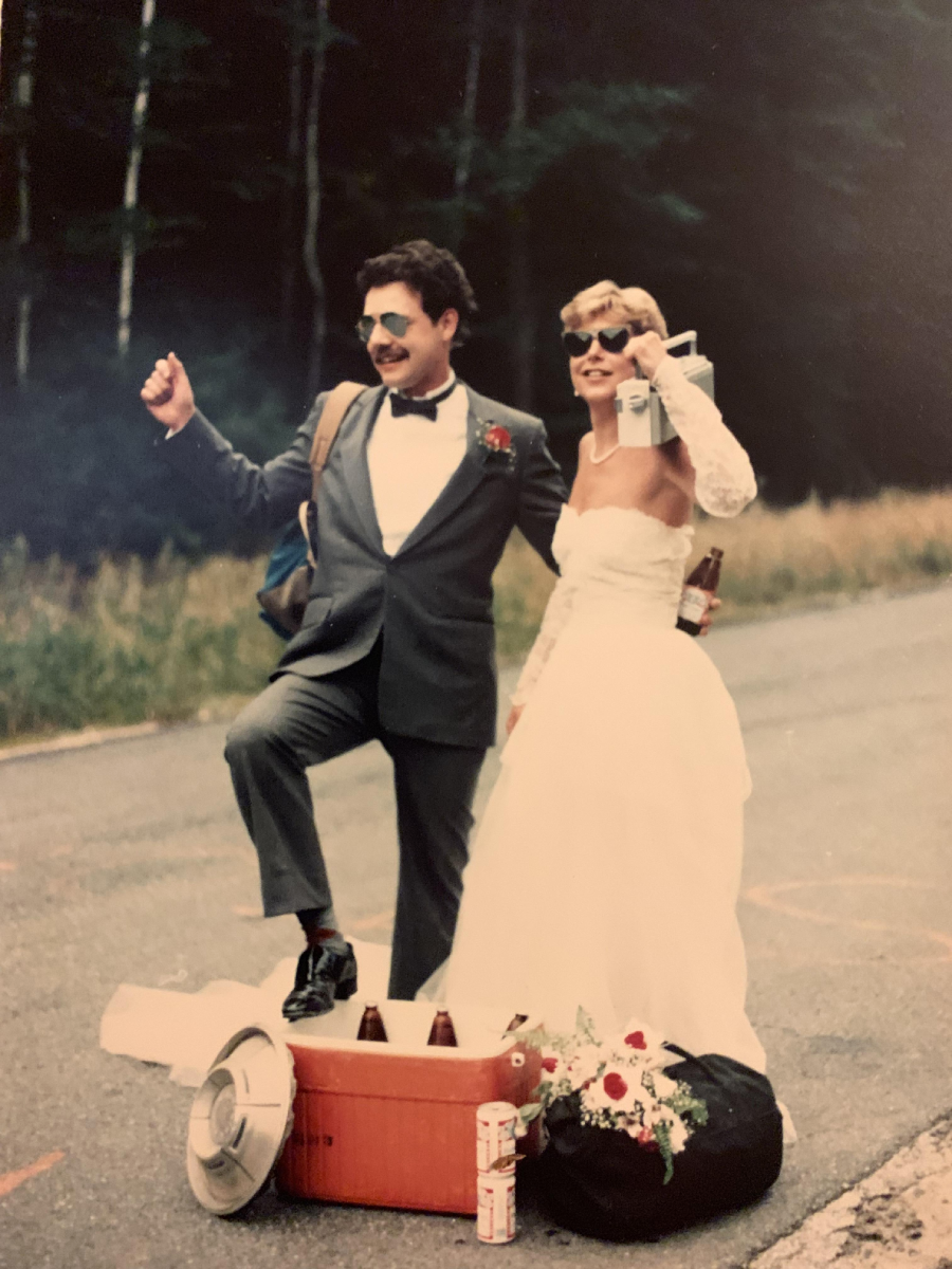 A bride and groom in wedding attire stand on a road, smiling and posing with drinks. They’re next to a cooler filled with bottles, a black bag, and a bouquet, with a forested background. The scene looks fun and informal.