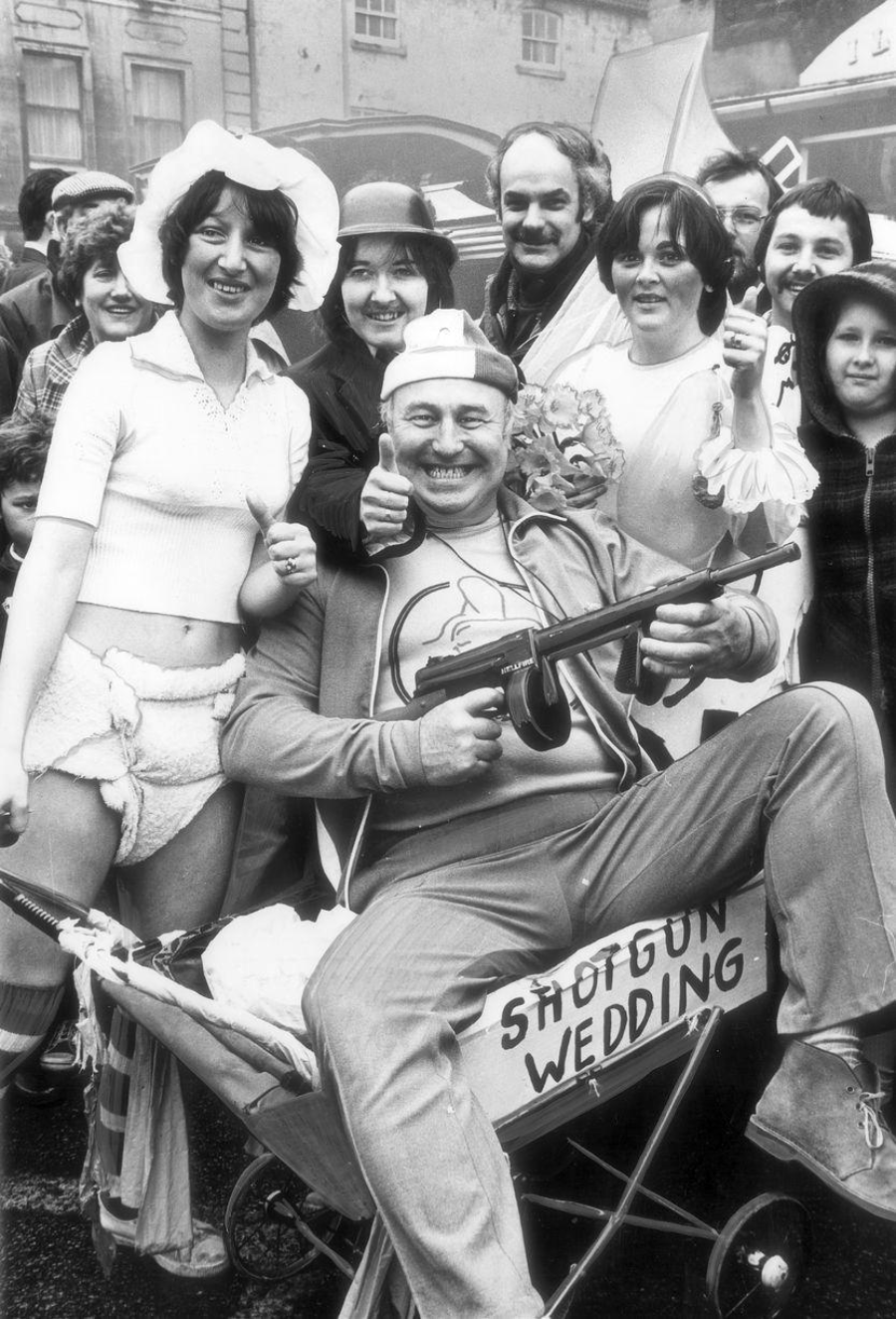 A group of people in humorous costumes pose for a photo. One person sits in a wheelbarrow labeled "SHOTGUN WEDDING," holding a toy gun, while others stand around them smiling, dressed in playful wedding and baby attire.