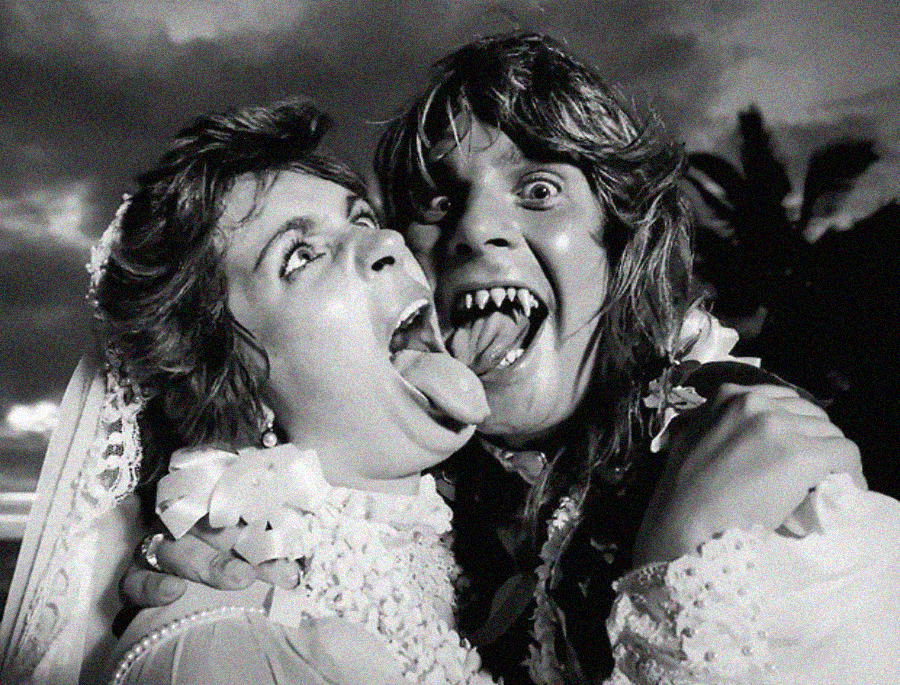 A black and white photo of two people in wedding attire playfully making faces; one sticks out their tongue while the other opens their mouth wide, baring vampire-like fangs.