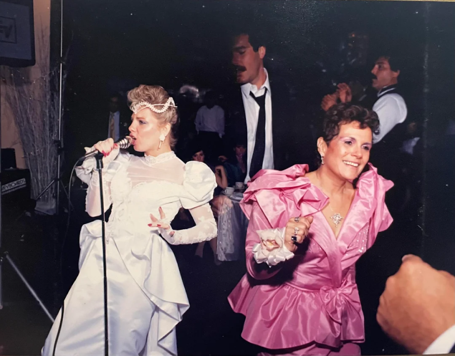 Two women in elaborate 1980s-style dresses, one in white singing into a microphone and one in pink dancing, are at a lively indoor party. Several people in suits are visible in the background.