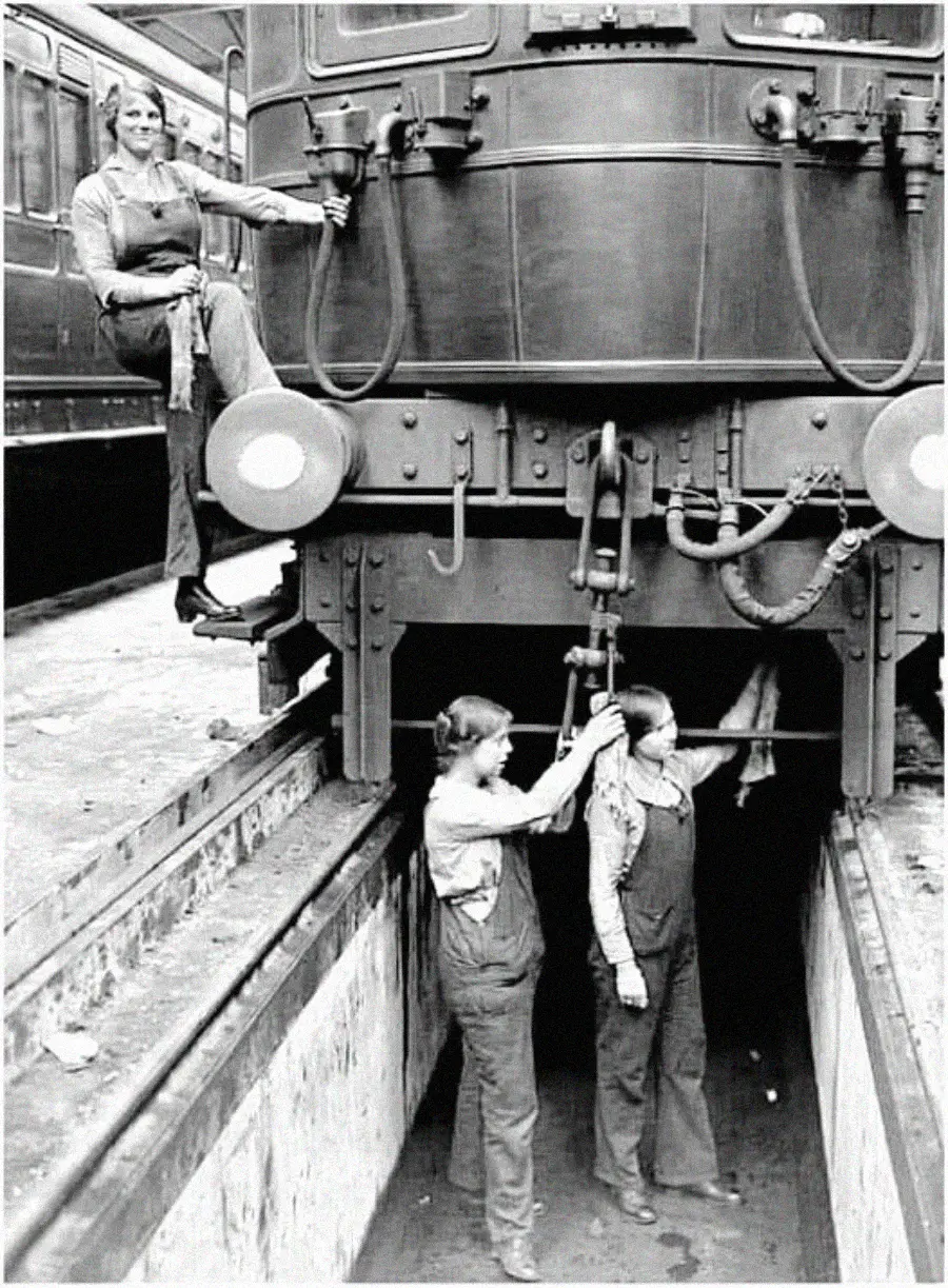 Three women in overalls work on maintaining a train; one stands smiling and holding a wrench on the front, while two others work beneath the train in a maintenance pit. The image is in black and white.