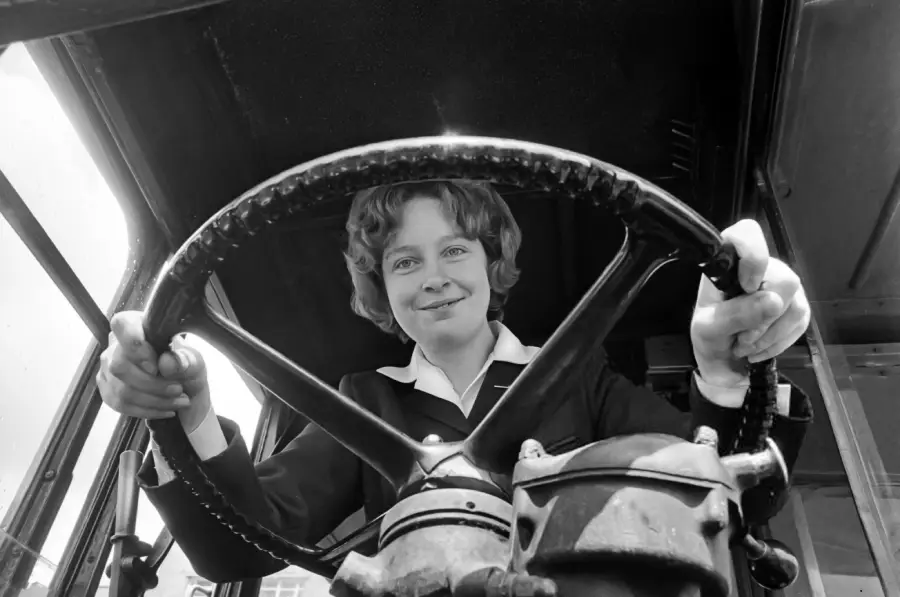 A young girl smiling as she sits behind the large steering wheel of a vehicle, possibly a bus or truck, viewed from a low angle inside the driver's seat.