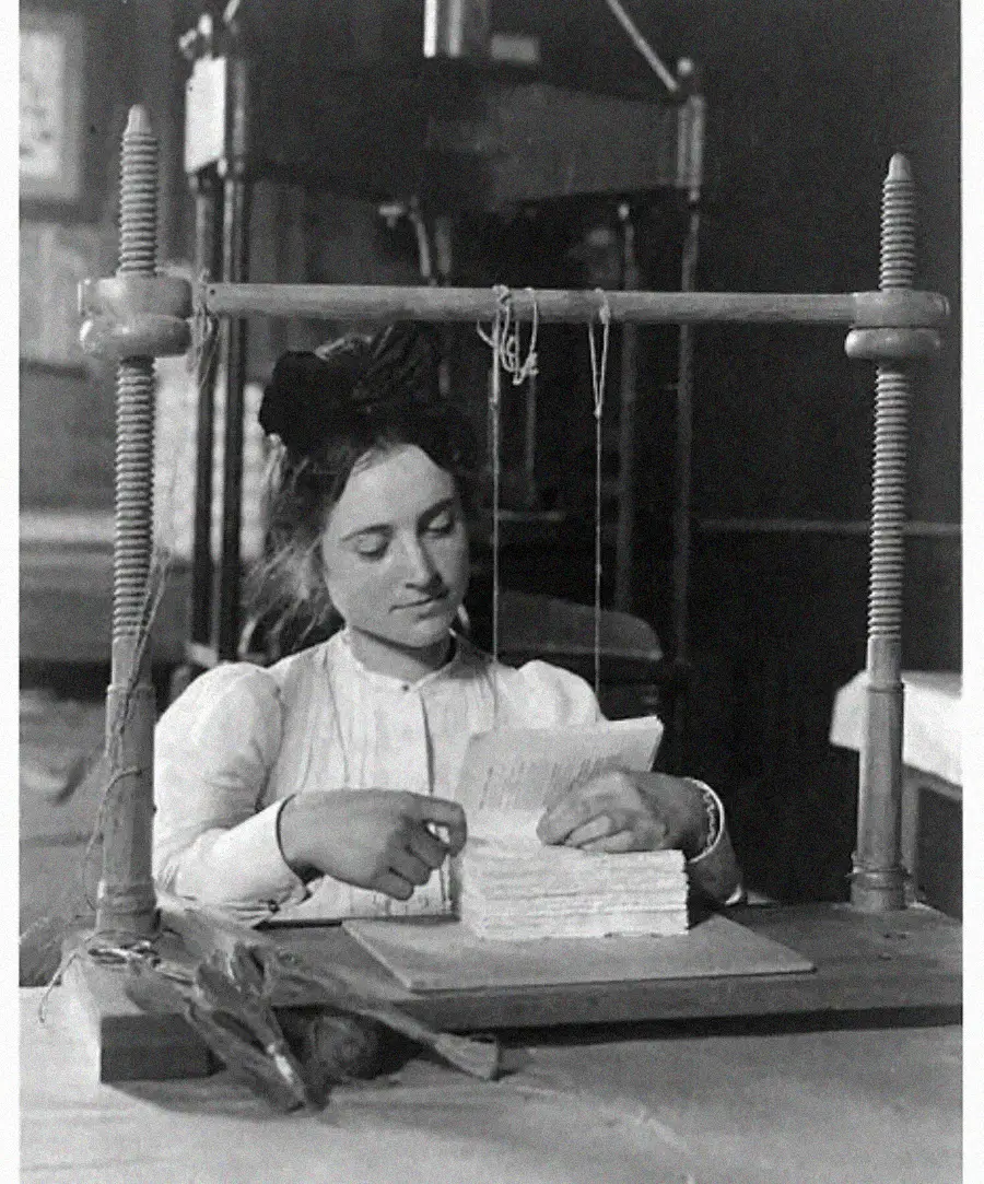 A woman in a long-sleeved blouse binds a stack of papers using a vintage bookbinding press, carefully sewing the pages together at a worktable.