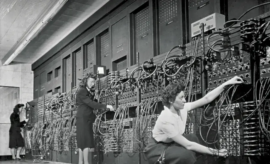 Three women work on a large, early computer system filled with cables and switches, plugging and unplugging wires into panels. The scene is in black and white, depicting mid-20th-century technology.