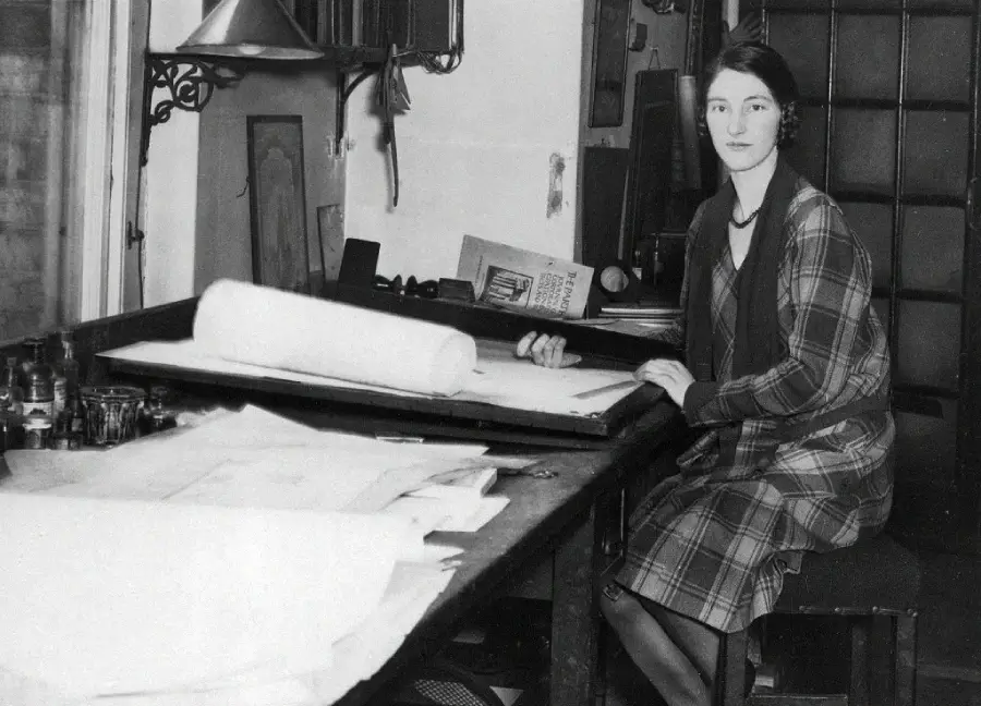 A woman in a plaid dress sits at a drafting table covered with papers and drawings, holding a rolled sheet. She looks at the camera, surrounded by work materials in a vintage office or studio setting.