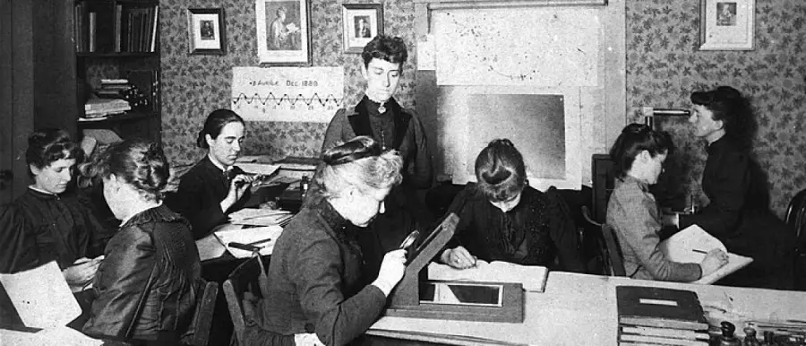 A group of women in Victorian-era clothing work and study at desks in a classroom, with papers, books, and instruments, as a teacher stands at the front. Framed pictures and patterned wallpaper decorate the room.
