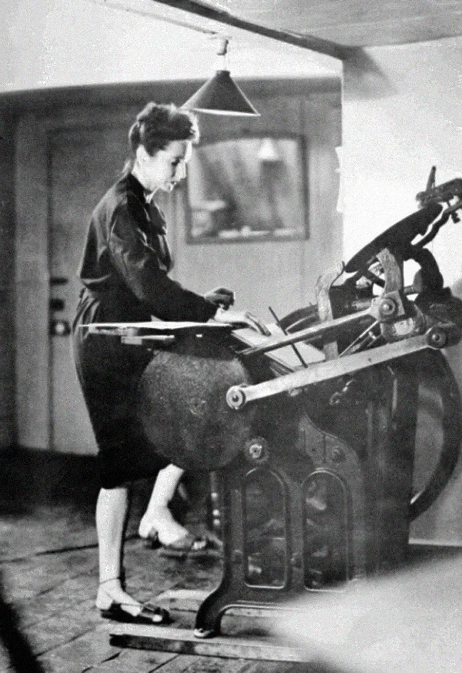 A woman stands operating an old-fashioned printing press in a room with wooden floors, concentrating on her work as she turns the machine's handle.