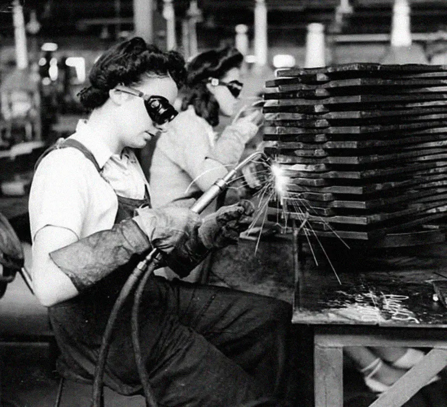 Two women wearing protective gloves and goggles are welding metal parts at a factory workstation; sparks fly from the welding torch as they work side by side, focused on their tasks.