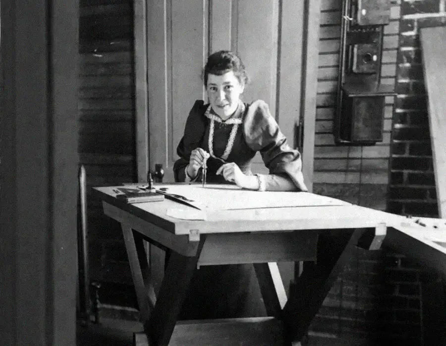A woman in a Victorian-era dress sits smiling at a large drafting table, holding a compass. The room has wooden walls, drawing tools, and technical equipment in the background.