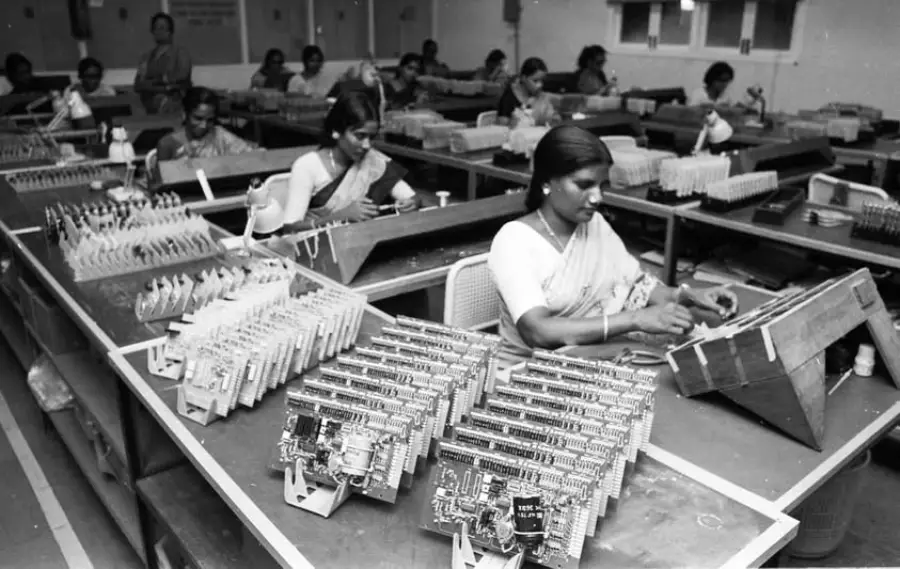 Women work at desks assembling electronic circuit boards in a factory setting. The room is filled with rows of components and workstations, and the workers wear traditional Indian attire.