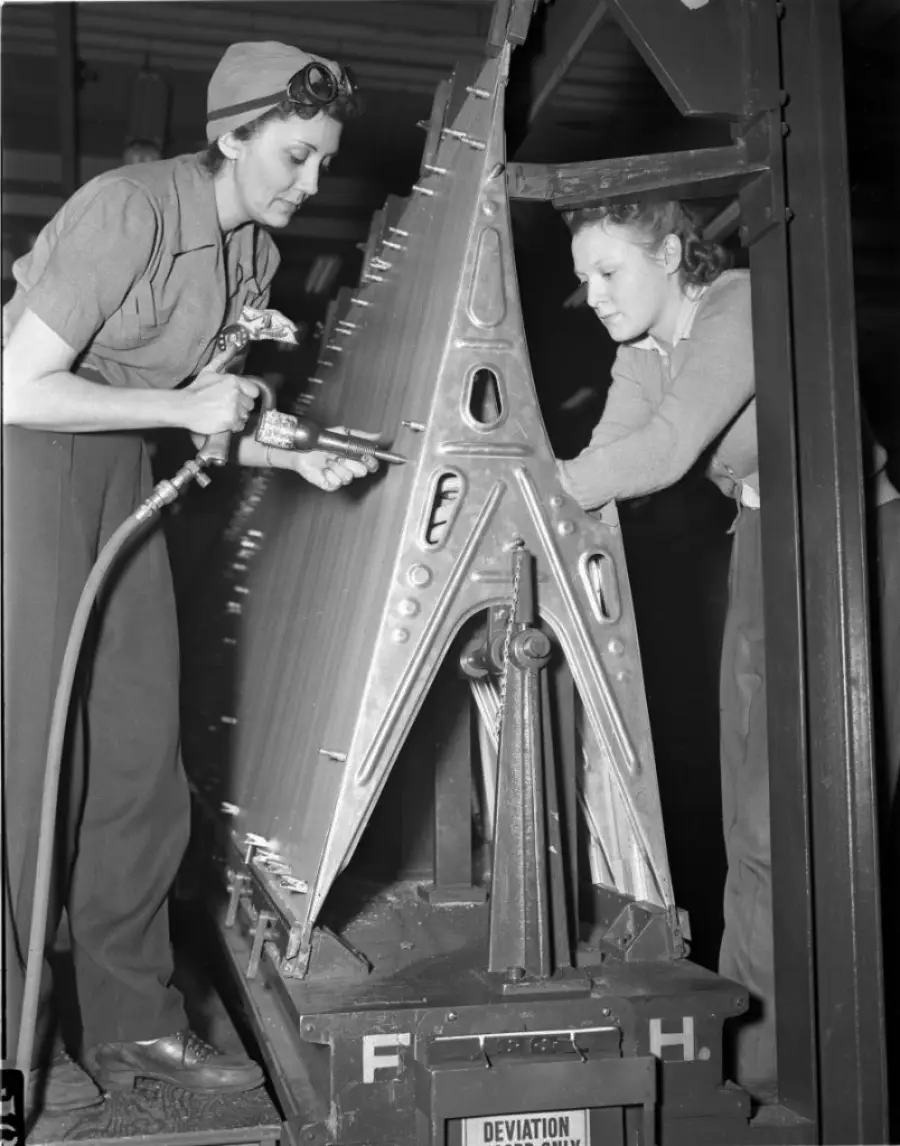 Two women, dressed in work clothes, use tools to assemble or rivet a large metal aircraft component in a factory setting during World War II. They focus intently on their work, highlighting women in industrial jobs.