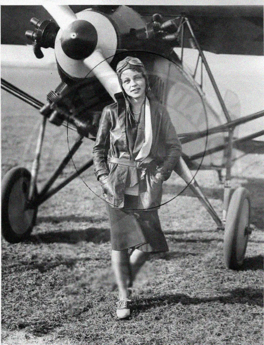 A woman in vintage aviator clothing stands in front of a propeller airplane on a grassy field, posing confidently with hands in her jacket pockets. The photo appears to be black and white and from the early 20th century.
