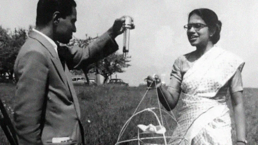 A man and a woman stand outdoors; the man holds a scientific instrument, while the woman, wearing a sari, holds a wire frame object. Both appear engaged in discussion, with trees and a car visible in the background.