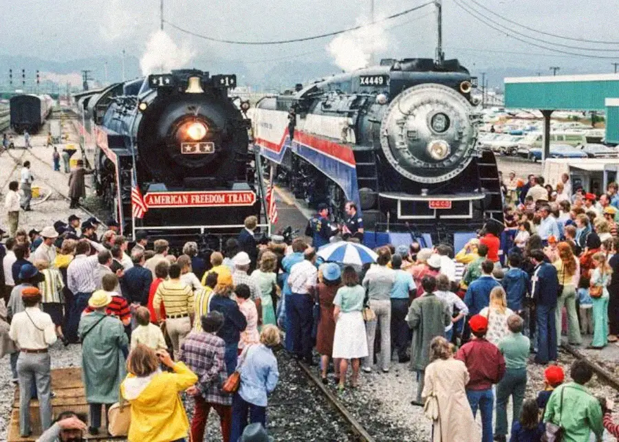 Two steam locomotives, including one labeled "American Freedom Train," are surrounded by a large crowd of people at a train station on a cloudy day. Many people are taking photos and looking at the trains.