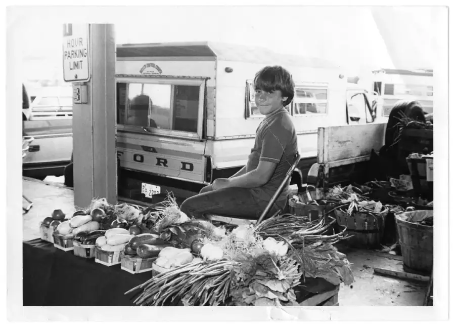 A young boy sits on a chair at a market stall with baskets of fresh vegetables in front of him. Behind him, a vintage pickup truck and other vehicles are parked. The scene appears to be outdoors, possibly at a farmer's market.