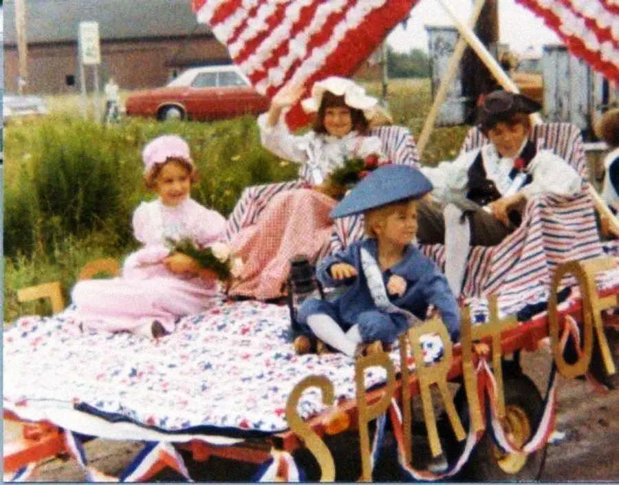 Four children in historical costumes sit on a parade float decorated with American flags and red, white, and blue fabric. The word "SPIRIT" is visible on the front. A red car and a building are in the background.