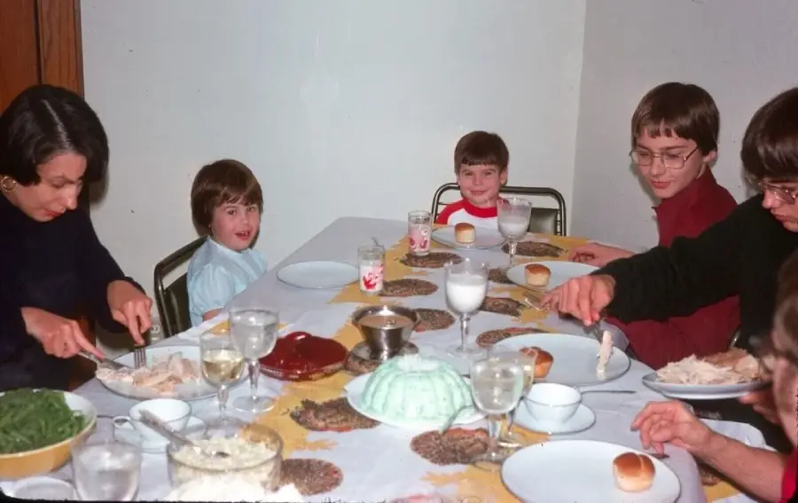 A family of six sits around a dining table set with food, drinks, and plates. Two children smile at the camera while adults serve food. The table is covered with a yellow and white tablecloth.
