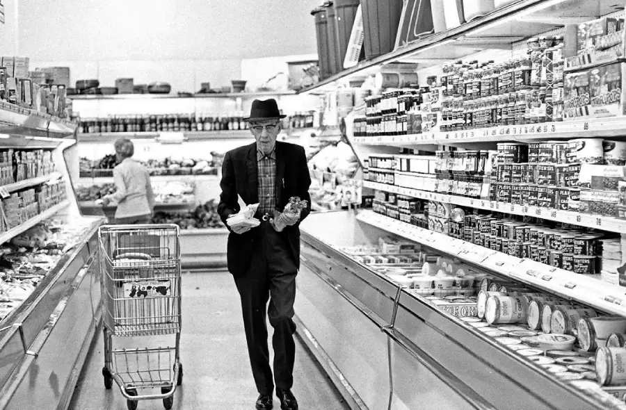 An elderly man wearing a hat and suit pushes a shopping cart down a grocery store aisle, holding groceries in his hands. Shelves are stocked with canned and packaged goods. Another person shops in the background.