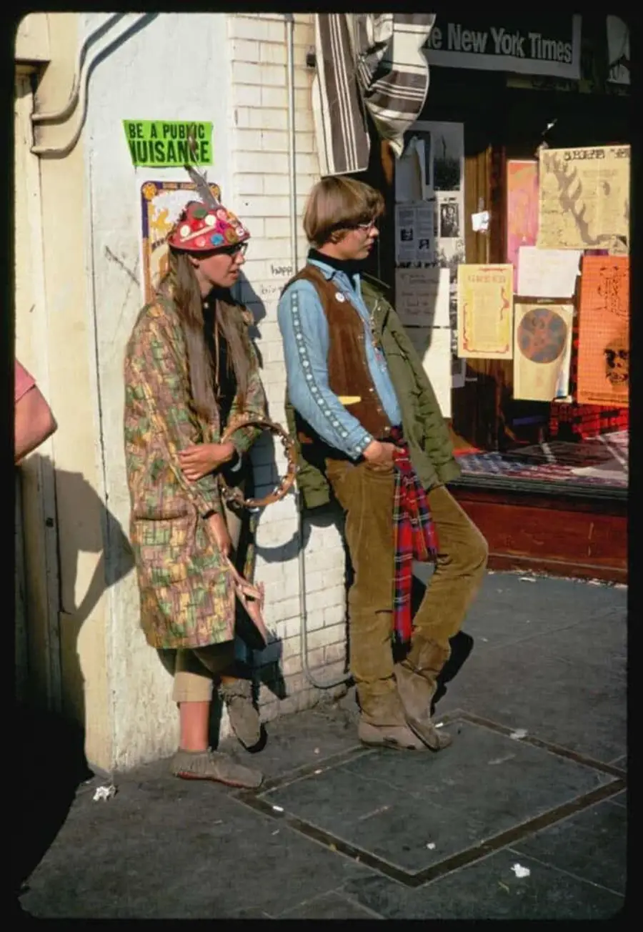 Two young people in colorful, bohemian clothes and accessories stand against a building wall on a city street, surrounded by posters, flyers, and a “Be a Public Nuisance” sign.