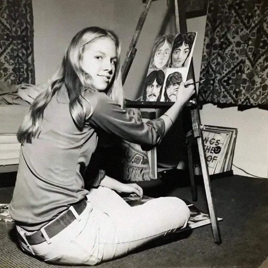 A young woman sits on the floor, painting portraits of four people on a canvas set on an easel in a cozy room with patterned curtains and framed artwork on the floor.