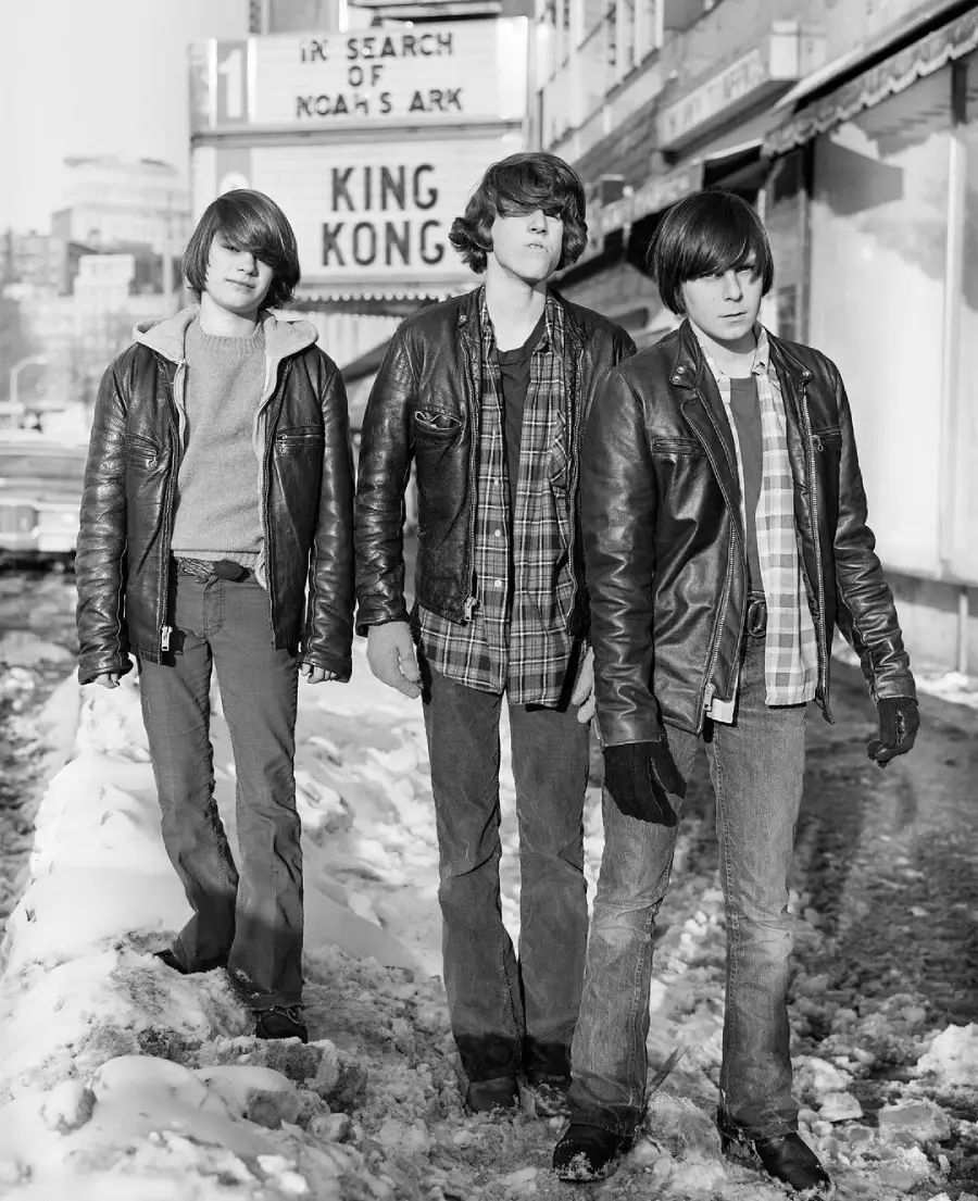 Three teenage boys in leather jackets stand on a snowy sidewalk in front of a movie theater showing "King Kong" and "In Search of Noah's Ark." The street and buildings are visible in the background.