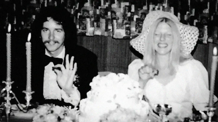 A black-and-white photo shows a man in a tuxedo and a woman in a white dress and wide-brimmed hat, sitting at a table with a cake, smiling and making hand gestures, possibly at a wedding reception.