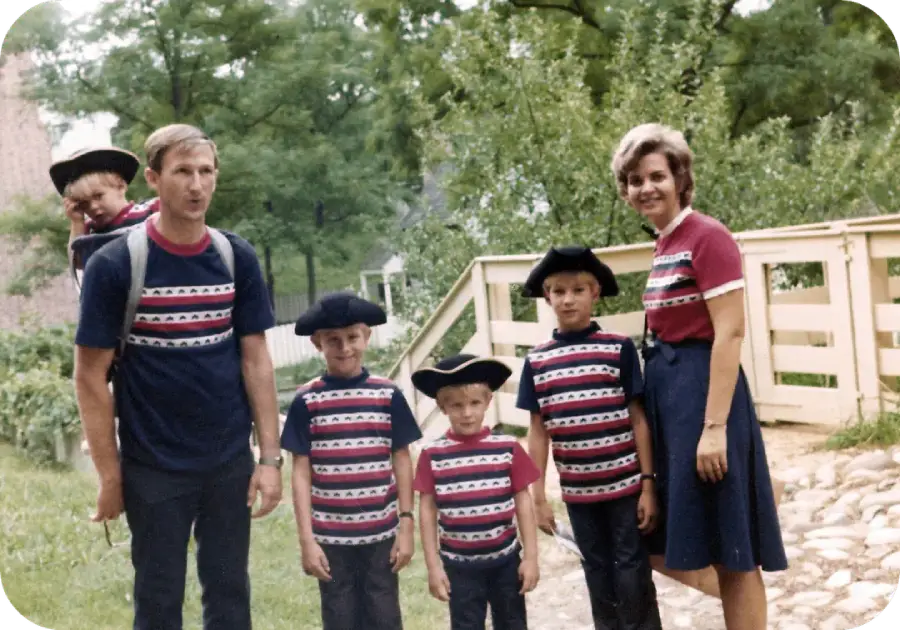 A family of six, all wearing matching red, white, and blue patterned shirts and dark bottoms, poses outdoors by a wooden bridge. The father carries a young child on his back; the three boys wear black hats.