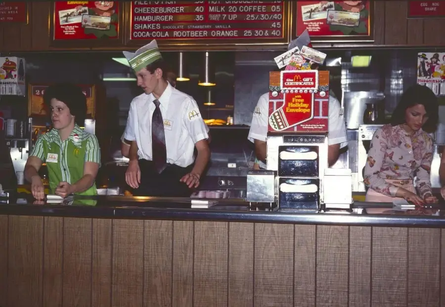 Three fast food workers stand behind a counter in a retro-style restaurant, with menus displayed above them; one wears a green uniform, another a white shirt with a tie, and the third is using a register.