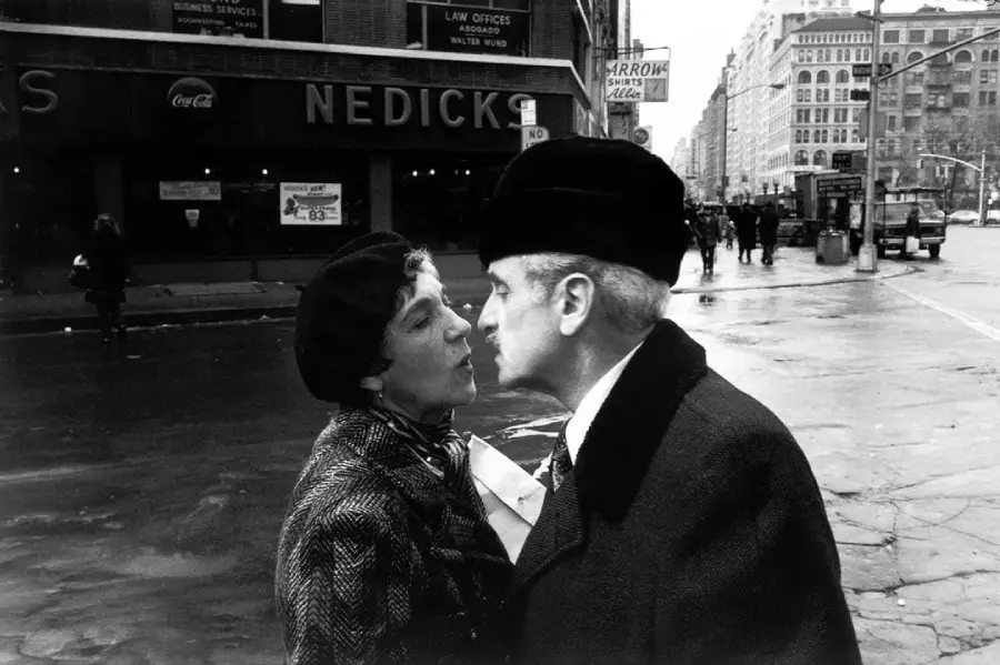 An older woman and man, both wearing dark hats and coats, lean toward each other on a city street, possibly about to kiss. Buildings, storefronts, and pedestrians are visible in the background.