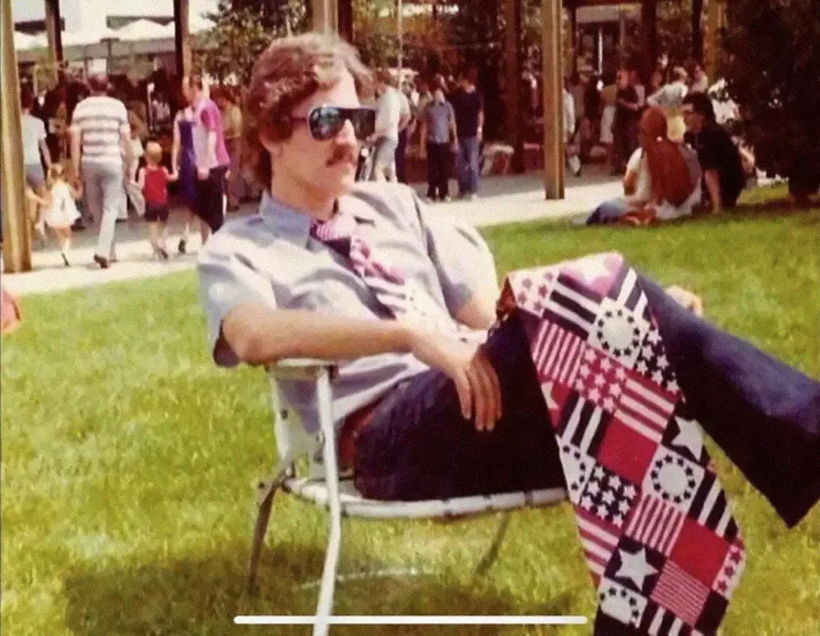 A man with sunglasses and a mustache relaxes in a lawn chair, wearing a patterned tie and distinctive flared pants with a patchwork of stars and stripes. People and trees are visible in the background on a sunny day.