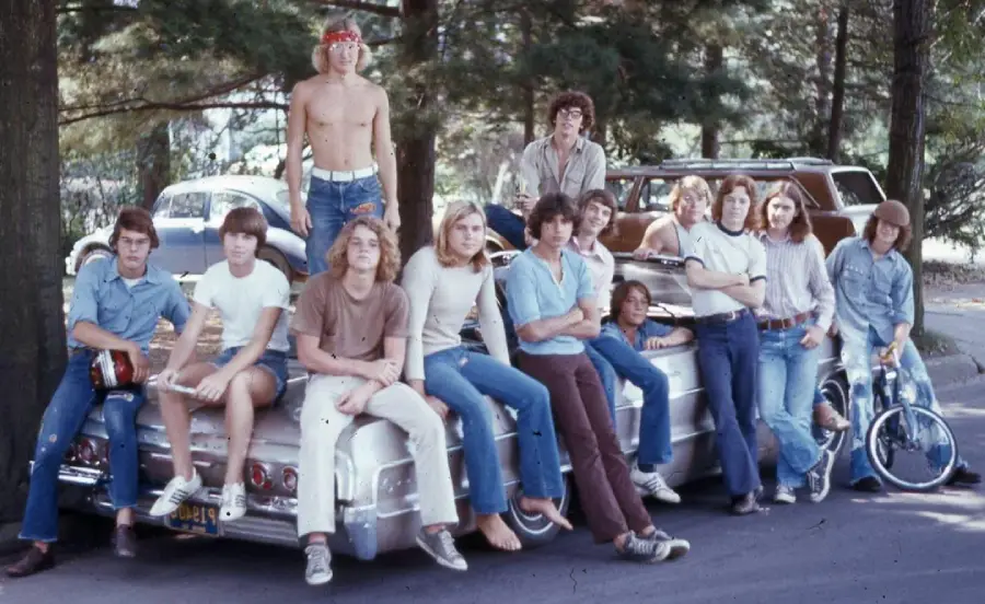A group of teenage boys in casual 1970s clothes pose on and around a classic car parked on a tree-lined street, with one shirtless boy standing on the trunk and others sitting or leaning against the car.