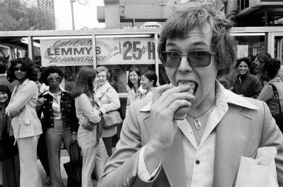A man in a suit and glasses eats a hot dog in the foreground while a group of smiling women stand in line behind him. A sign in the background reads “Celebrate Lemmy’s 25th Anniversary.”