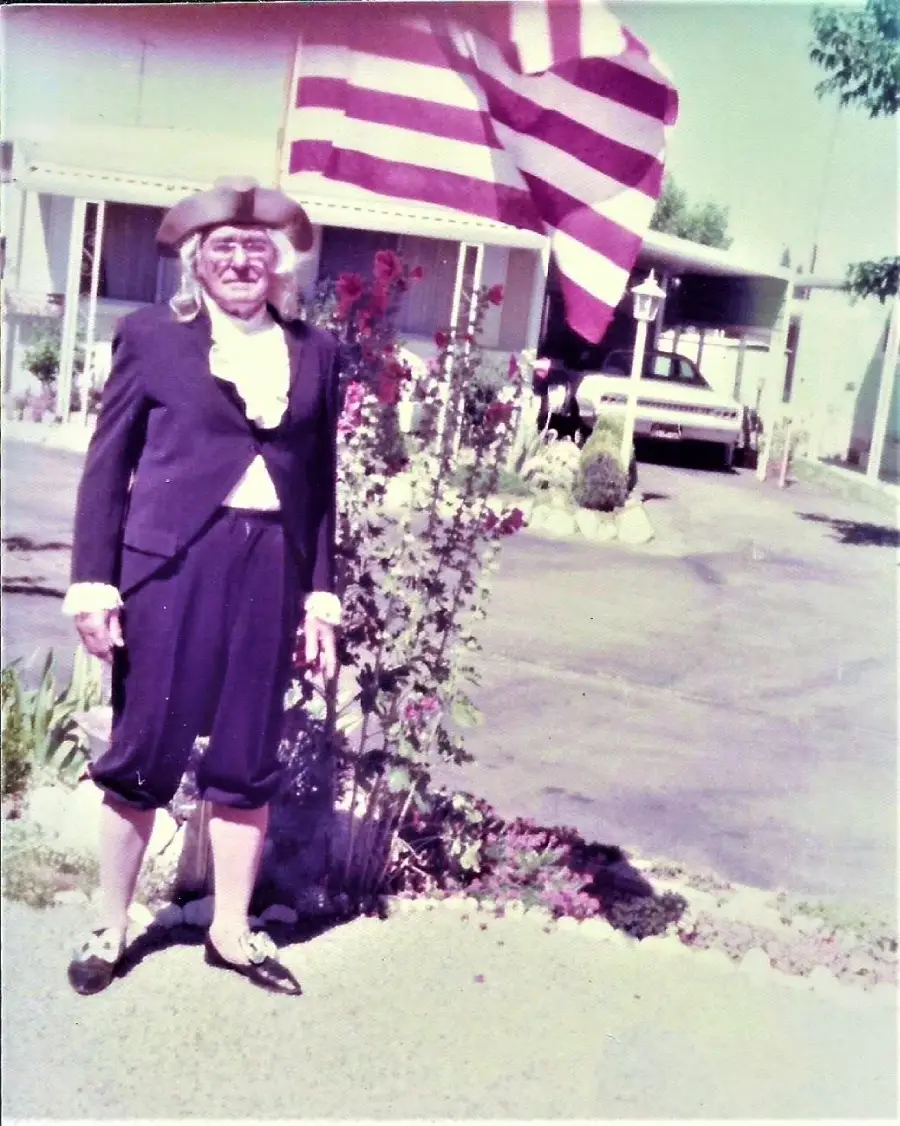 An older person dressed in colonial-era clothing stands outside near a large American flag, with a carport and garden visible in the background on a sunny day.