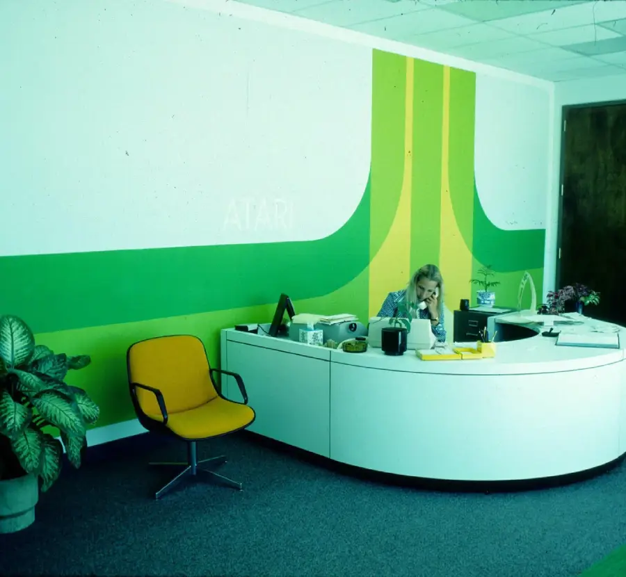 A woman sits at a curved white reception desk in a vintage office with a green and yellow Atari logo mural on the wall, near an orange chair and potted plant.