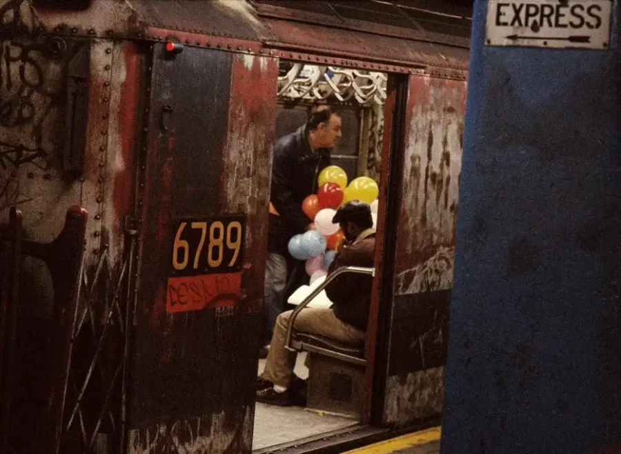 A man stands holding colorful balloons near a seated person inside a graffiti-covered subway car numbered 6789, with part of a blue column labeled "EXPRESS" visible on the platform.