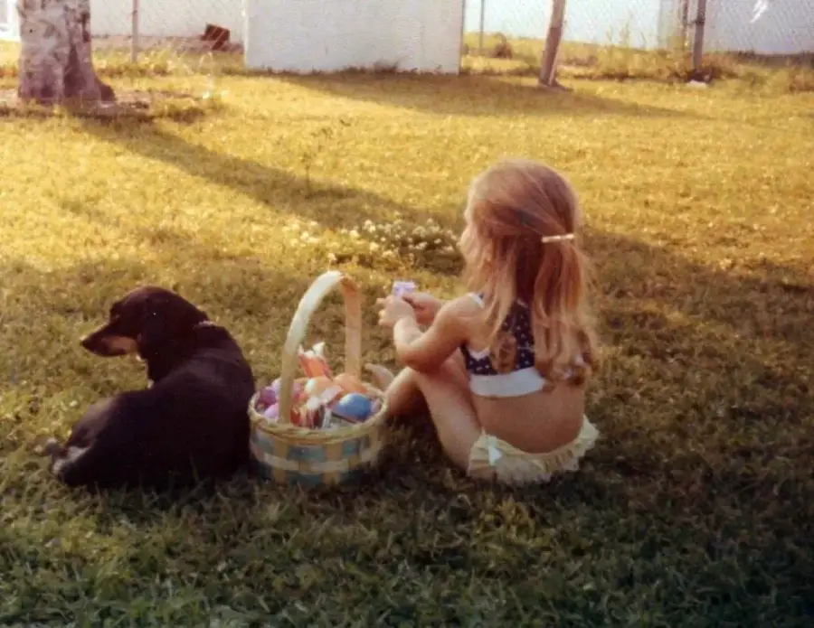 A young girl in a bikini sits on grass next to a black dog and an Easter basket filled with eggs, outdoors on a sunny day with a fence and trees in the background.