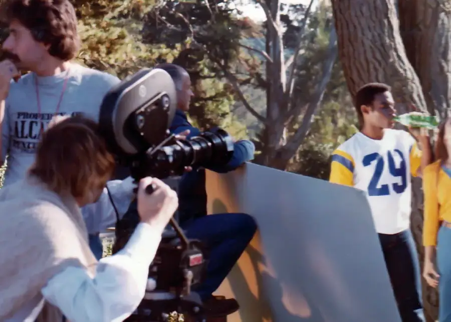 A film crew shoots a scene outdoors with a camera and reflector, while a man in a blue and yellow number 29 jersey drinks from a green bottle near a woman in a matching yellow top. Trees are visible in the background.