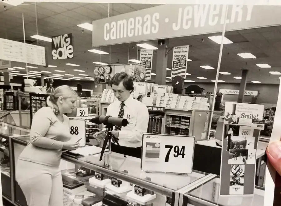 A woman and a man stand at a glass counter under a "Cameras • Jewelry" sign in a store. The man shows her a telescope while the woman holds a box. Price signs and shelves of products are visible in the background.