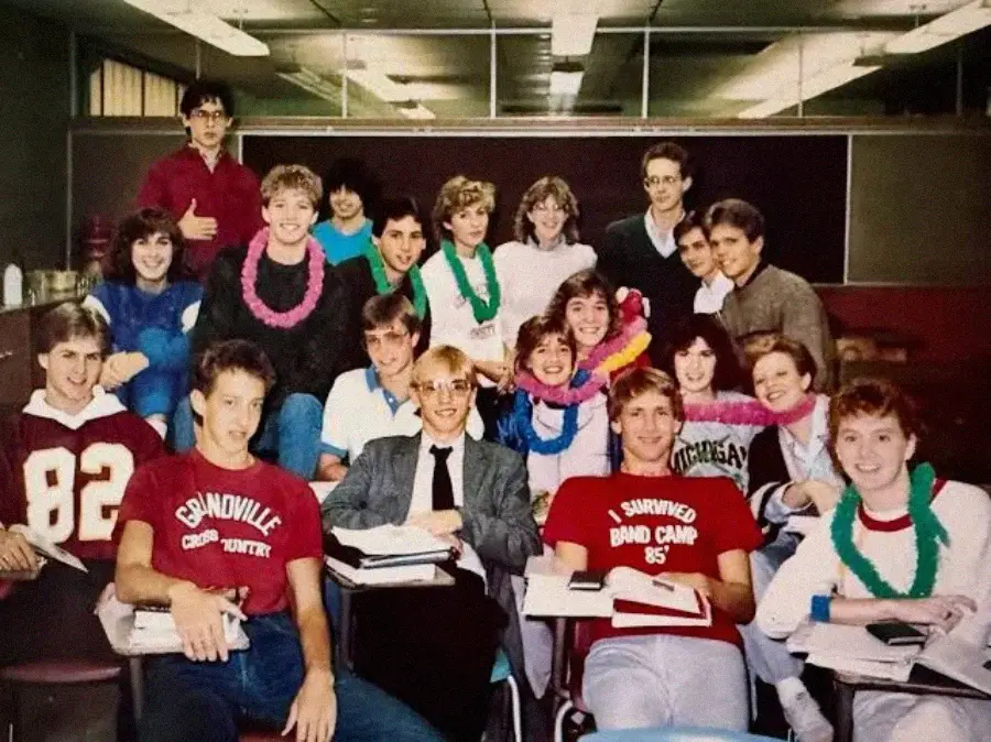 A group of smiling teenagers pose together in a classroom, some wearing colorful leis. They are sitting and standing around desks with books and notebooks, appearing cheerful and relaxed.